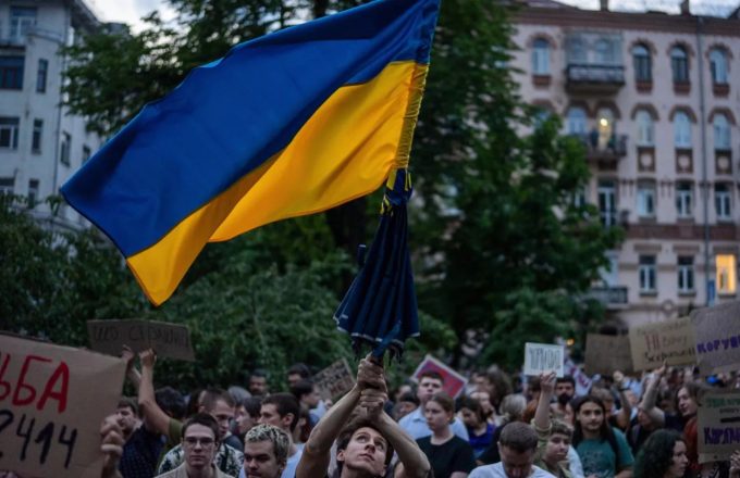 A crowd of protesters walking, with one of them holding up a large Ukrainian flag