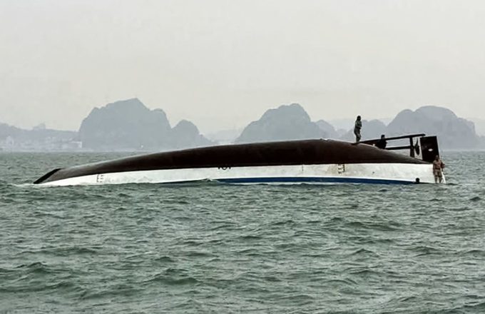 A capsized boat with some people trying to climb onto the hull to avoid drowning