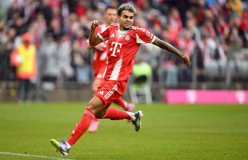 Luis Díaz running across the field in Bayern’s striking new red uniform while looking happy