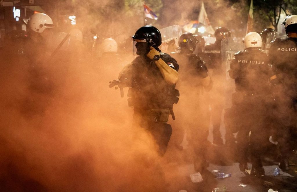 A heavily armed police officer in a black helmet and uniform, emerging dramatically from a cloud of tear gas
