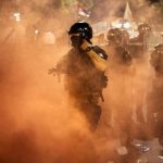 A heavily armed police officer in a black helmet and uniform, emerging dramatically from a cloud of tear gas