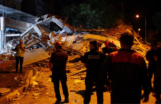 A completely destroyed structure with some rescuers walking over it searching for injured people