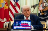 Donald Trump sitting in his presidential office with many flags behind him
