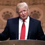 The President of the United States standing at a podium with arms outstretched as he addresses the crowd