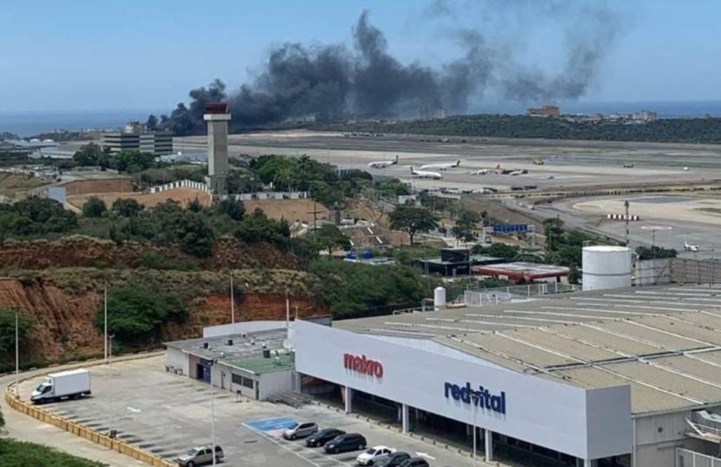 A large airport runway with a massive blaze in the distance caused by the private jet accident