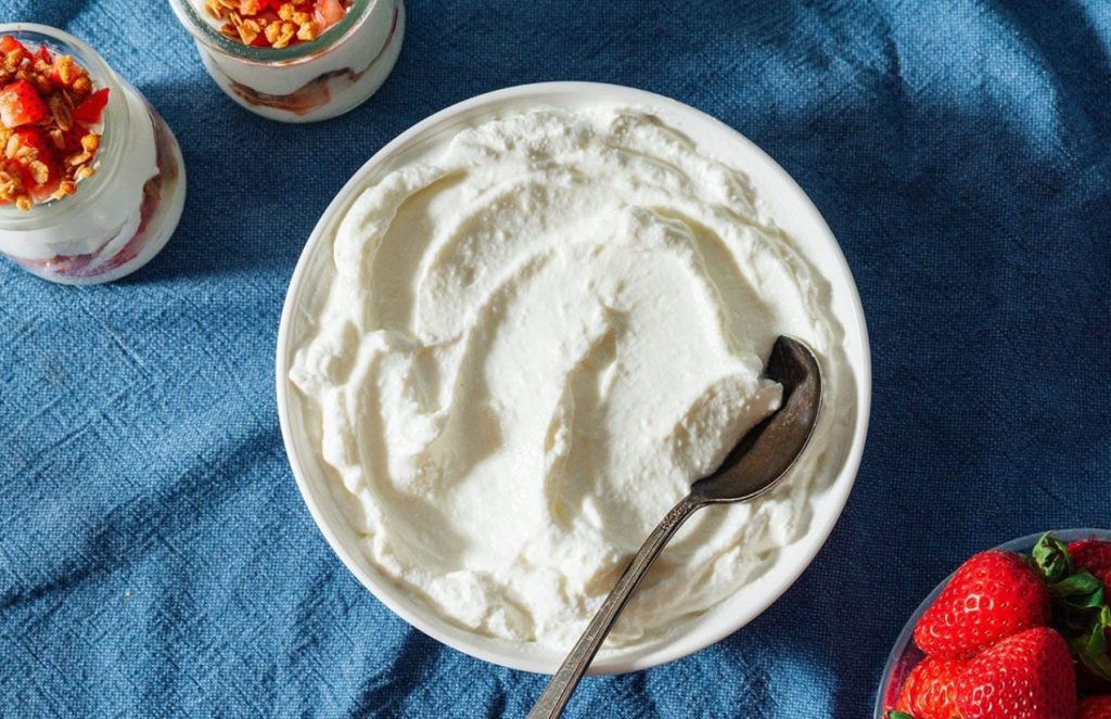 A large bowl filled with white Greek yogurt, with small jars of strawberries beside it