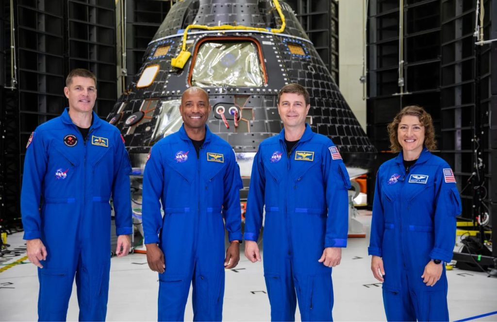 Four astronauts, including one woman, posing while standing in front of the head of the rocket that could take them into space