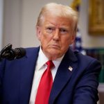 Trump wearing a suit, sitting at a desk with a stern expression