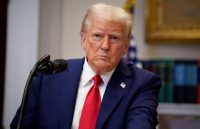 Trump wearing a suit, sitting at a desk with a stern expression
