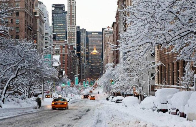 One of the streets of New York covered in snow, with a few people walking and some cars passing by