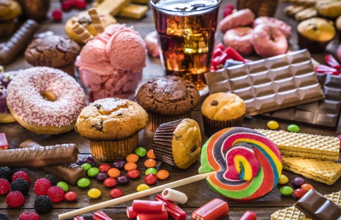 New York introduces mandatory added sugar warning on chain restaurant menus A large wooden table filled with sugary products such as Coca-Cola bottles, donuts, candies, puddings, and other sweets