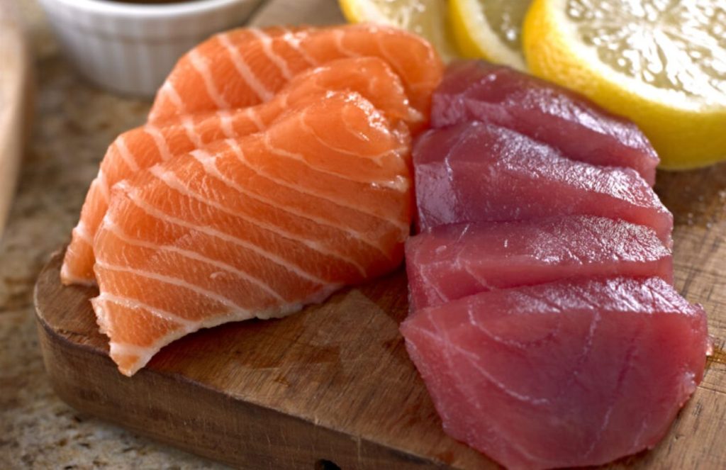 Three pieces of salmon and tuna on a cutting board accompanied by several slices of lemon