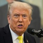 Trump sitting at his desk surrounded by multiple microphones as he speaks to reporters
