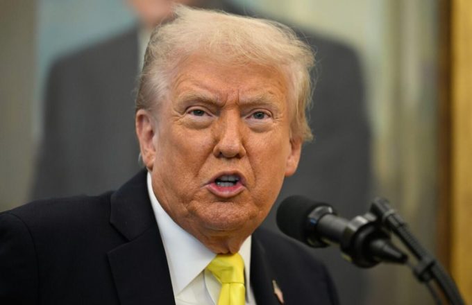 Trump sitting at his desk surrounded by multiple microphones as he speaks to reporters