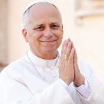 The Pope smiling with his hands together as he stands in front of his followers