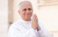 The Pope smiling with his hands together as he stands in front of his followers
