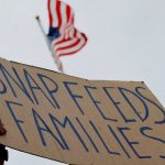 The hands of a person holding up a cardboard sign that reads “SNAP FEEDS FAMILIES,” with the flag of the United States in the background
