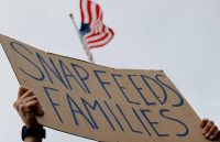 The hands of a person holding up a cardboard sign that reads “SNAP FEEDS FAMILIES,” with the flag of the United States in the background
