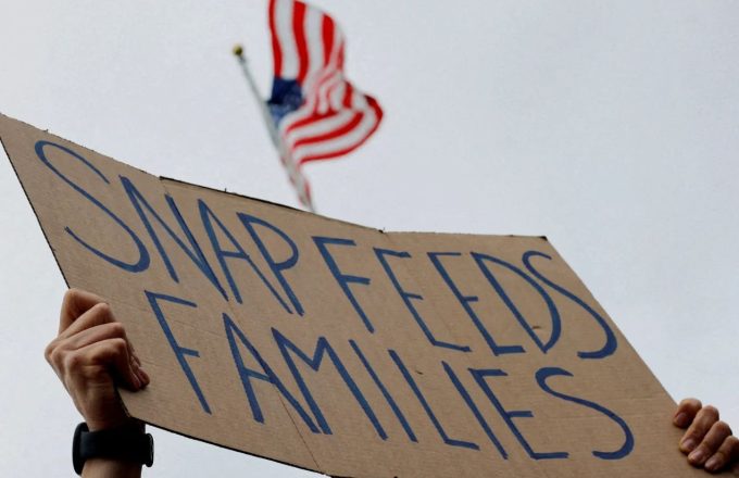 The hands of a person holding up a cardboard sign that reads “SNAP FEEDS FAMILIES,” with the flag of the United States in the background