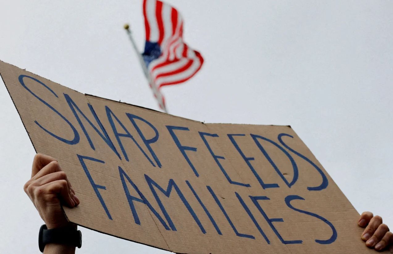 The hands of a person holding up a cardboard sign that reads “SNAP FEEDS FAMILIES,” with the flag of the United States in the background