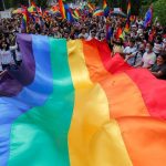 Many LGBTQ+ people holding one of the movement’s flags in the middle of a march