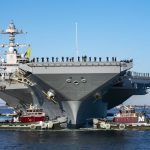 A front-view shot of the incredible warship as it is being guided by several smaller tugboats