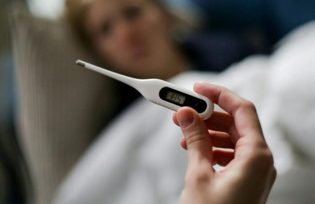 A person holding a thermometer showing a high temperature, with another person lying in bed in the background