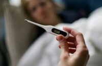 A person holding a thermometer showing a high temperature, with another person lying in bed in the background