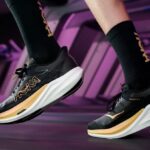 A model posing with the new black and gold HOKA shoes, as if they were running.