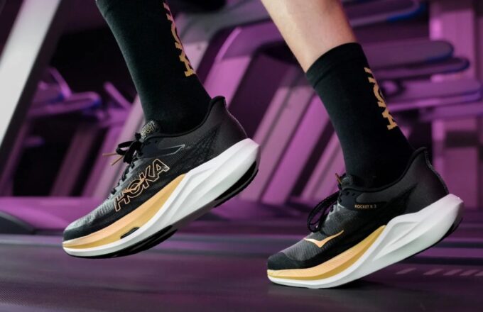 A model posing with the new black and gold HOKA shoes, as if they were running.