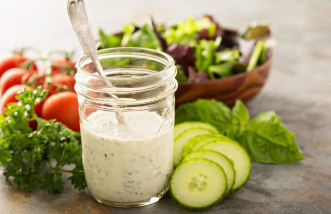 Several vegetables on a table alongside a glass of dressing with a spoon inside