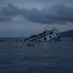 A ferry slowly sinking while its passengers, in the water, watch it go down