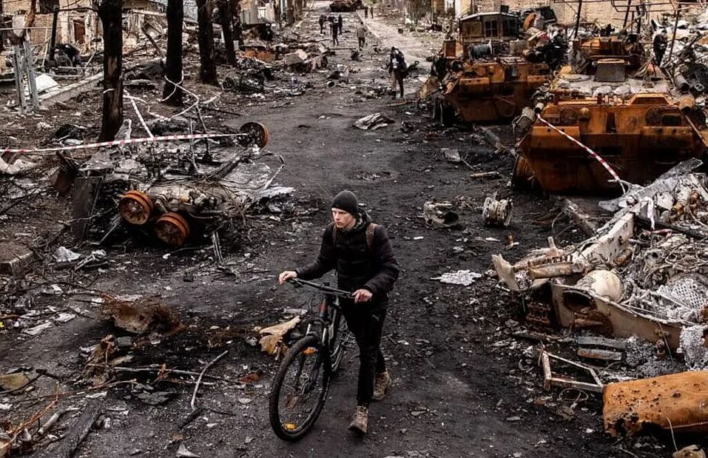 A person walking with a bicycle in hand through a city destroyed by war