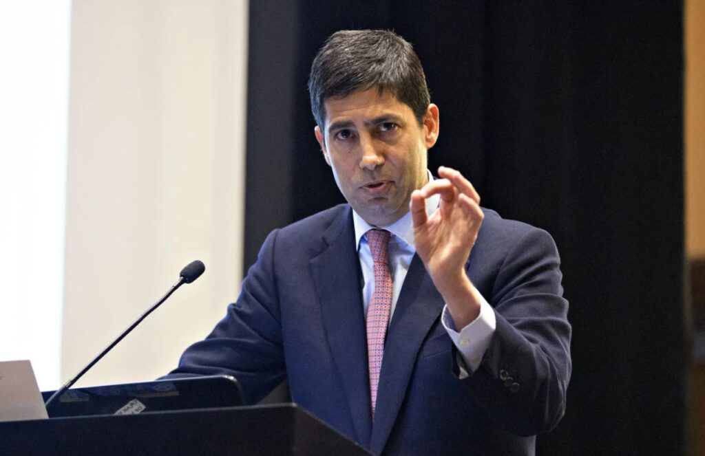 Kevin Warsh wearing an elegant suit, delivering a speech in front of a large audience