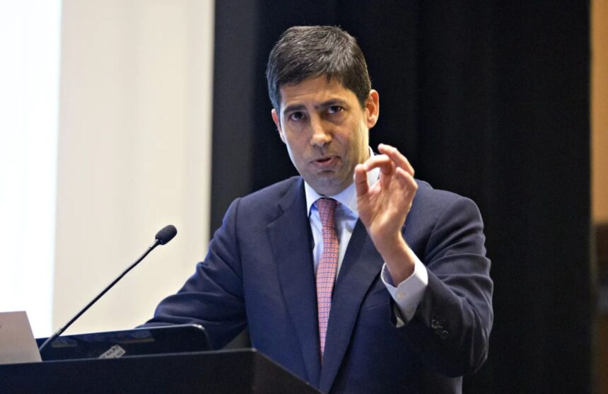 Kevin Warsh wearing an elegant suit, delivering a speech in front of a large audience