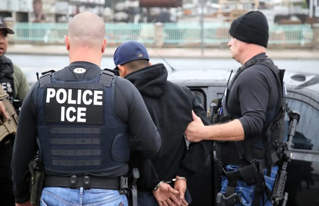 Federal judge halts immigration arrests in Oregon without a judicial warrant Several ICE agents, with one of them turning his back to the camera while wearing a vest that reads “ICE Police”