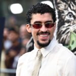 Bad Bunny posing for the cameras on a red carpet in an elegant and eye-catching white suit with shiny accessories