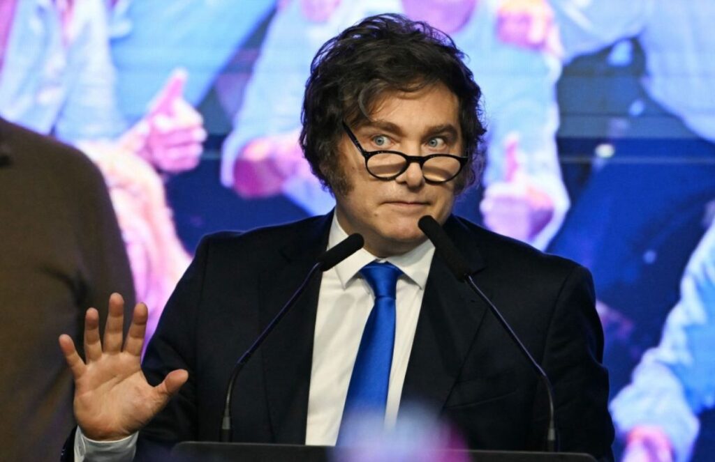 Milei wearing an elegant suit and glasses while speaking in front of his supporters and fellow political colleagues