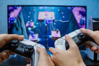 Two people holding PS5 controllers while playing on a large TV