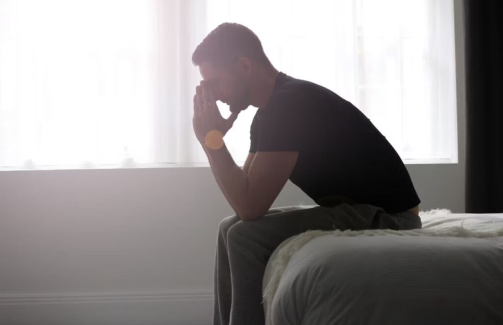 A silhouette of a man sitting on the edge of a bed with a sad posture