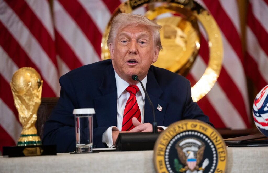Trump sitting in a meeting with a soccer ball and the World Cup trophy beside him