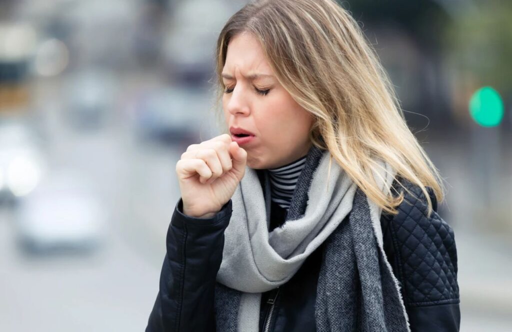 A woman holding her chest while coughing outdoors