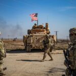 Several soldiers walking with their equipment while being escorted by a tank