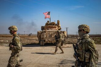 Several soldiers walking with their equipment while being escorted by a tank