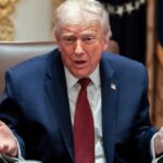 Donald Trump sitting at a desk with his hands open while explaining something to someone