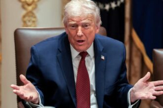 Donald Trump sitting at a desk with his hands open while explaining something to someone