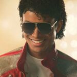 Michael Jackson smiling at his audience wearing his iconic red jacket and large sunglasses.
