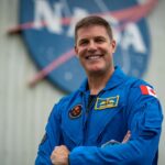 A photo of a space pilot smiling while wearing a spacesuit, with the NASA logo in the background