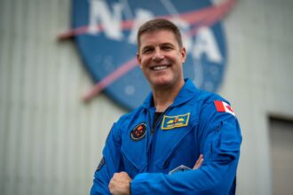 A photo of a space pilot smiling while wearing a spacesuit, with the NASA logo in the background