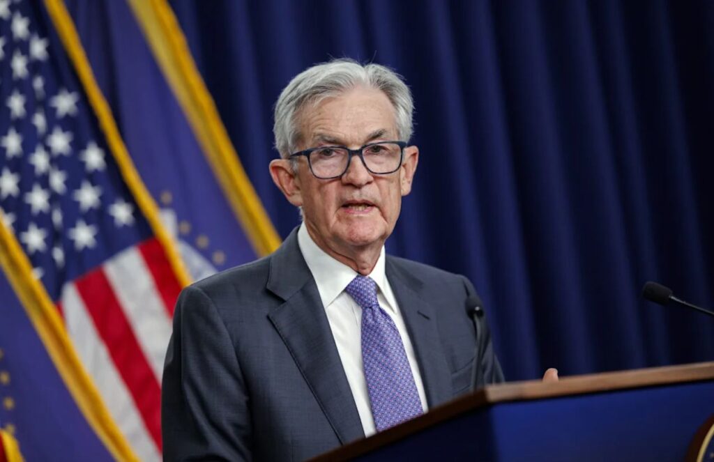 Jerome Powell standing at a podium giving a press conference in front of many cameras and journalists.